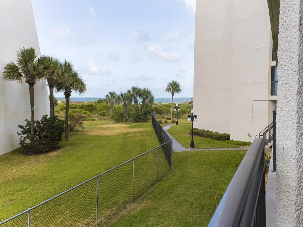 View from balcony overlooking green lawn, palm trees, and sandy beach with ocean horizon beyond.