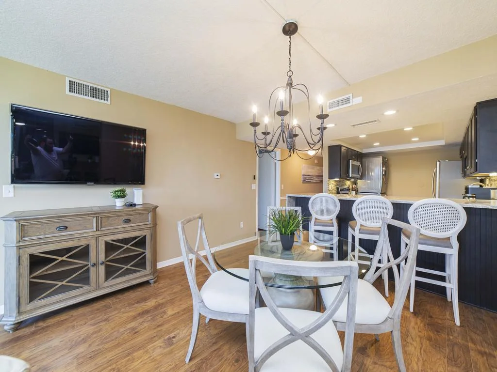 Dining area with round glass table, cushioned wood chairs, and wrought iron chandelier over hardwood floors.