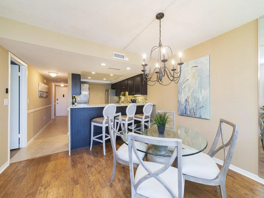 Glass-top dining table with cushioned cross-back chairs under a wrought iron chandelier near the kitchen bar.