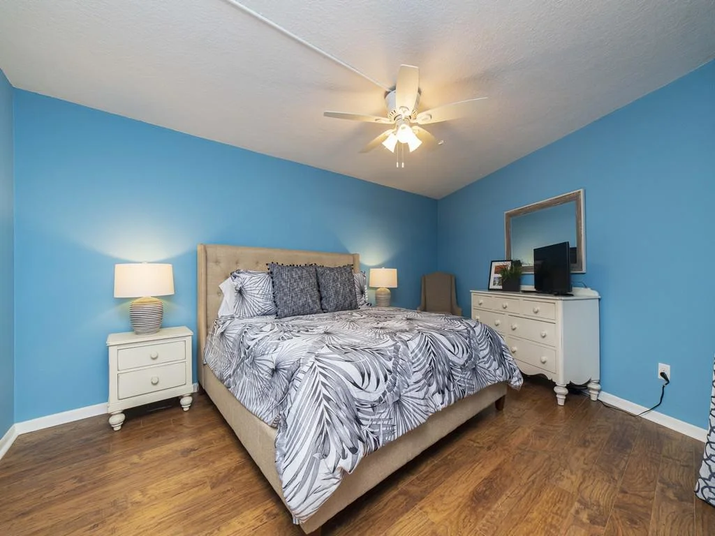 Second bedroom with blue walls, wood floors, beige upholstered bed, white dresser, and ceiling fan lighting.