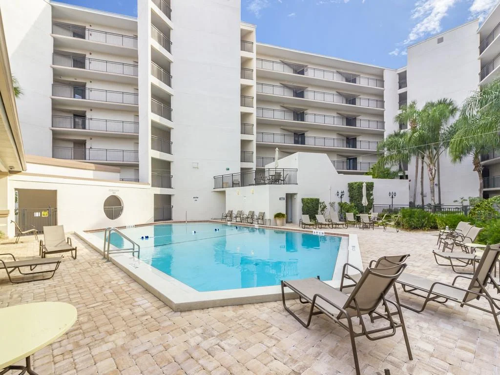Rectangular pool with beige stone pavers and metal lounge chairs set against white condo walls and palm trees.