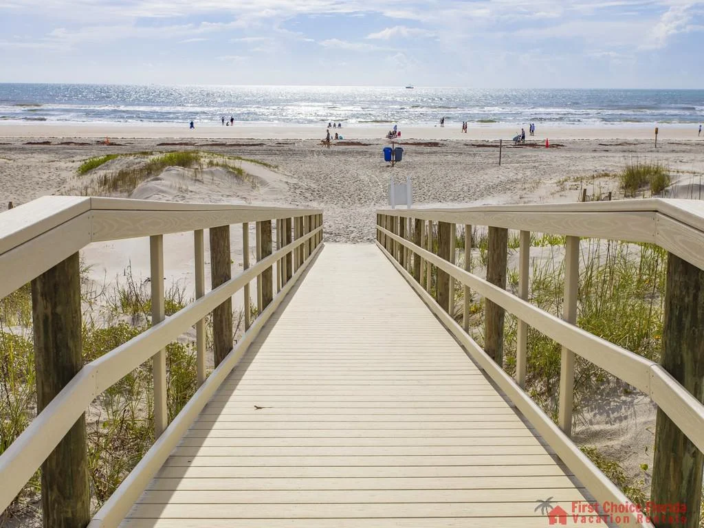 Wooden boardwalk with railings extends over beach dunes toward sunlit ocean waves and scattered beachgoers.
