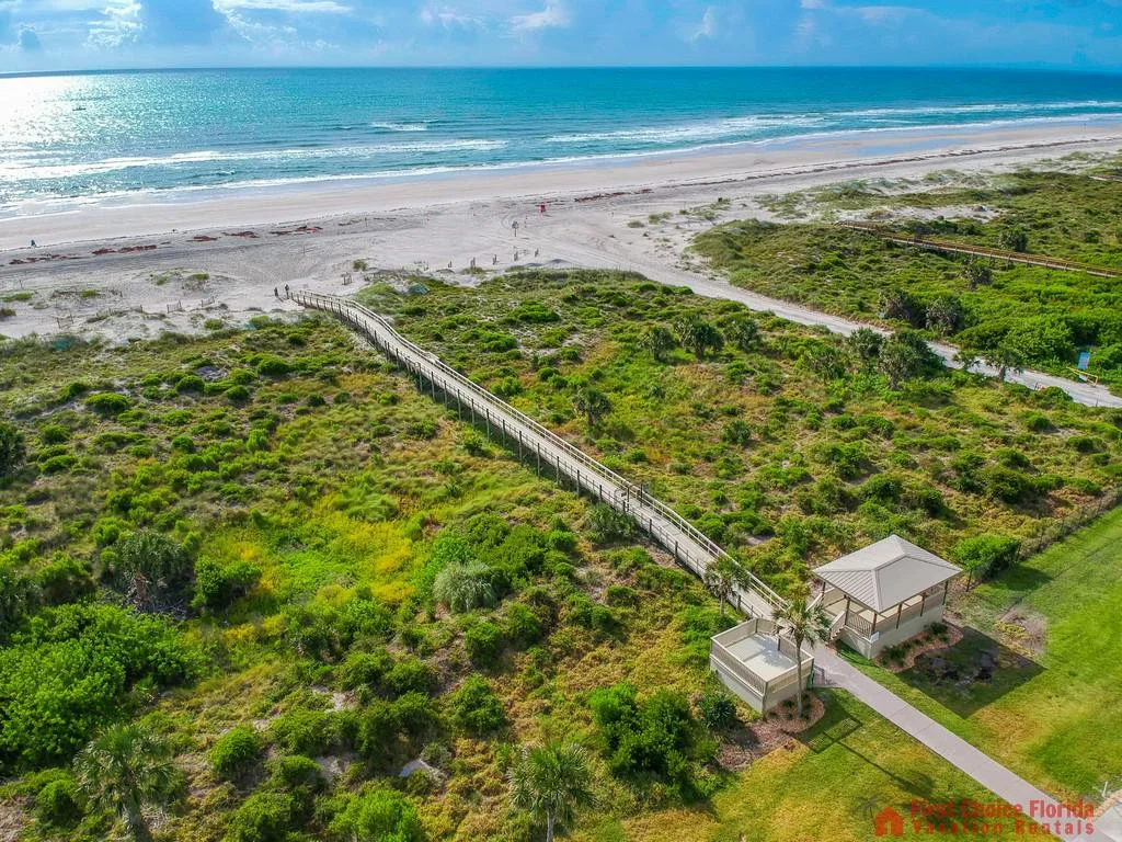 Boardwalk stretches over coastal greenery toward the sandy beach and shimmering ocean under a partly cloudy sky.
