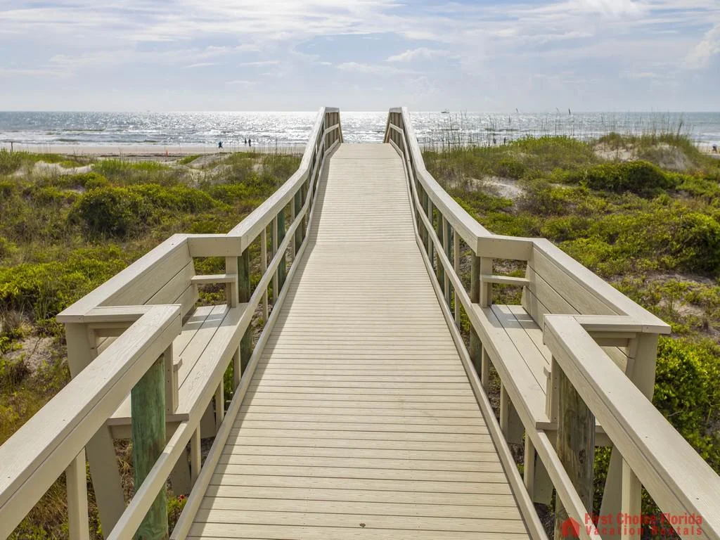 Wooden boardwalk with benches leads through dunes to the sunlit ocean shore under a partly cloudy sky.
