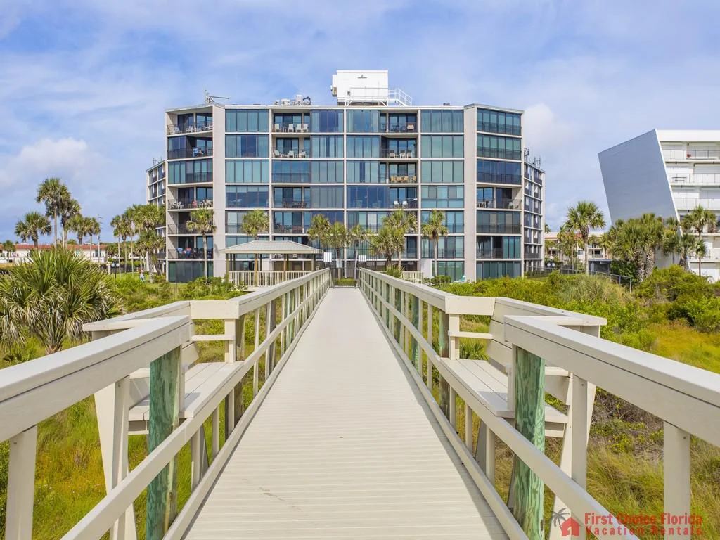 Boardwalk with white railings leads through coastal greenery to a seven-story glass-front condo building.