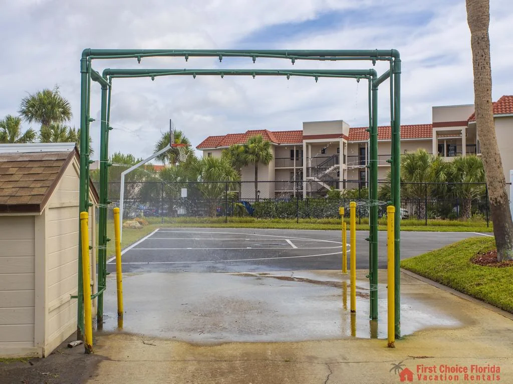 Outdoor shower area with green metal frame and yellow posts, overlooking a basketball half-court and palm trees.