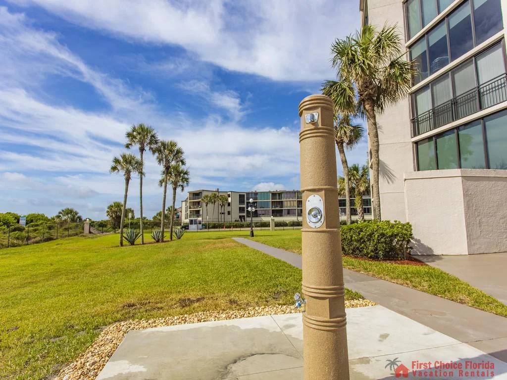 Outdoor beach shower station stands on a concrete pad next to a walkway, with palm trees and condos nearby.