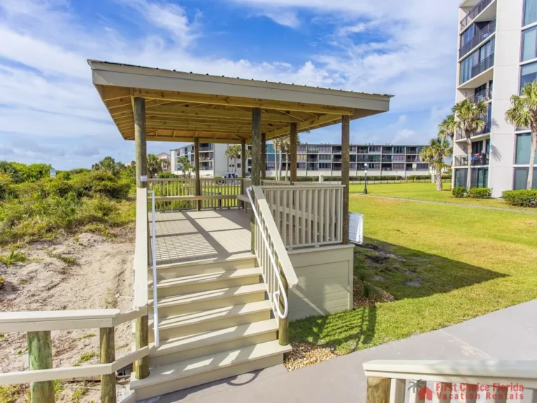Covered wooden gazebo with beige railings, stairs, and posts overlooks sandy dunes and nearby condo buildings.