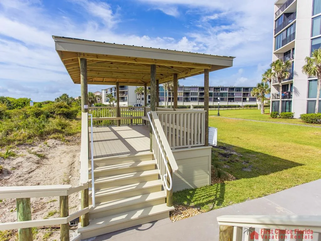 Covered wooden gazebo with beige railings, stairs, and posts overlooks sandy dunes and nearby condo buildings.