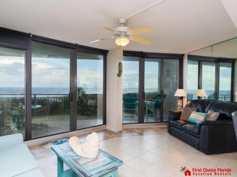 Floor-to-ceiling sliding glass doors open to an oceanfront balcony with black leather sofa and tiled floors.
