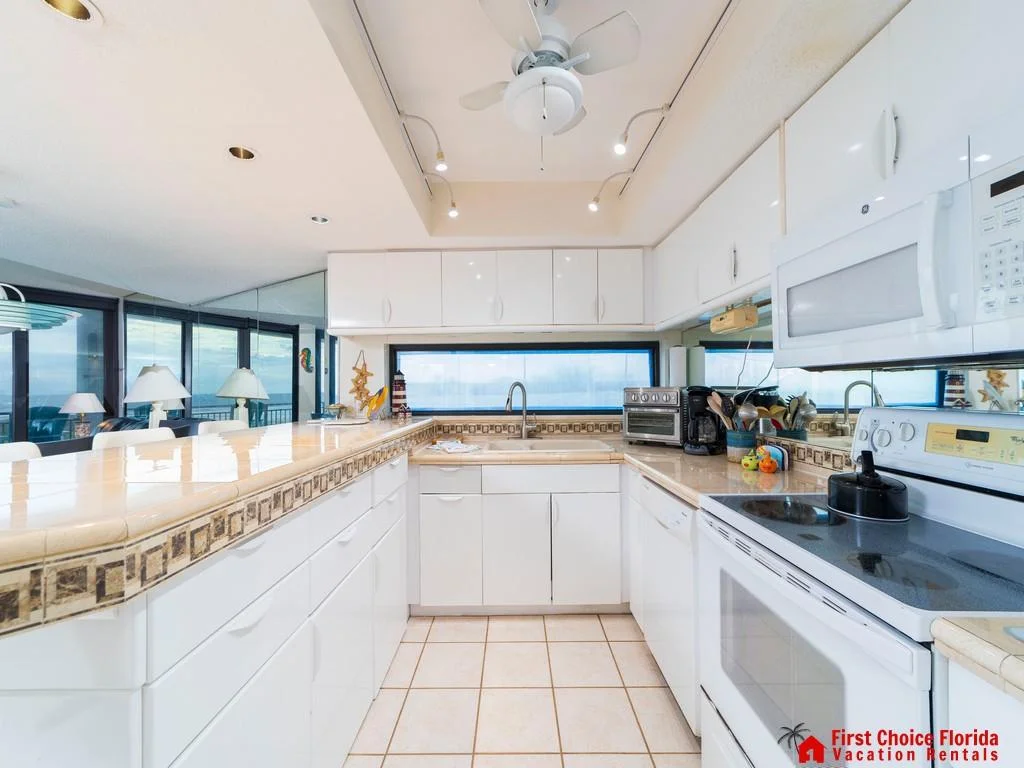 White cabinetry and tiled counters with decorative trim surround a corner sink beneath a wide window.