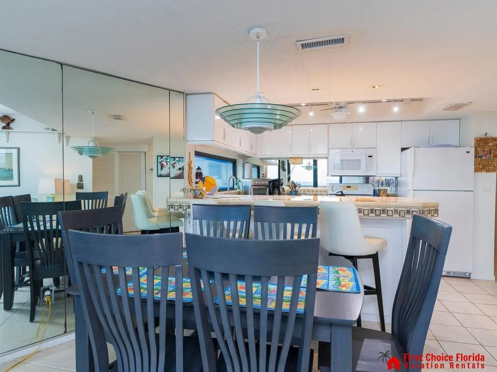 Dark wood dining table with colorful placemats sits beside a mirrored wall reflecting the kitchen’s white cabinetry.