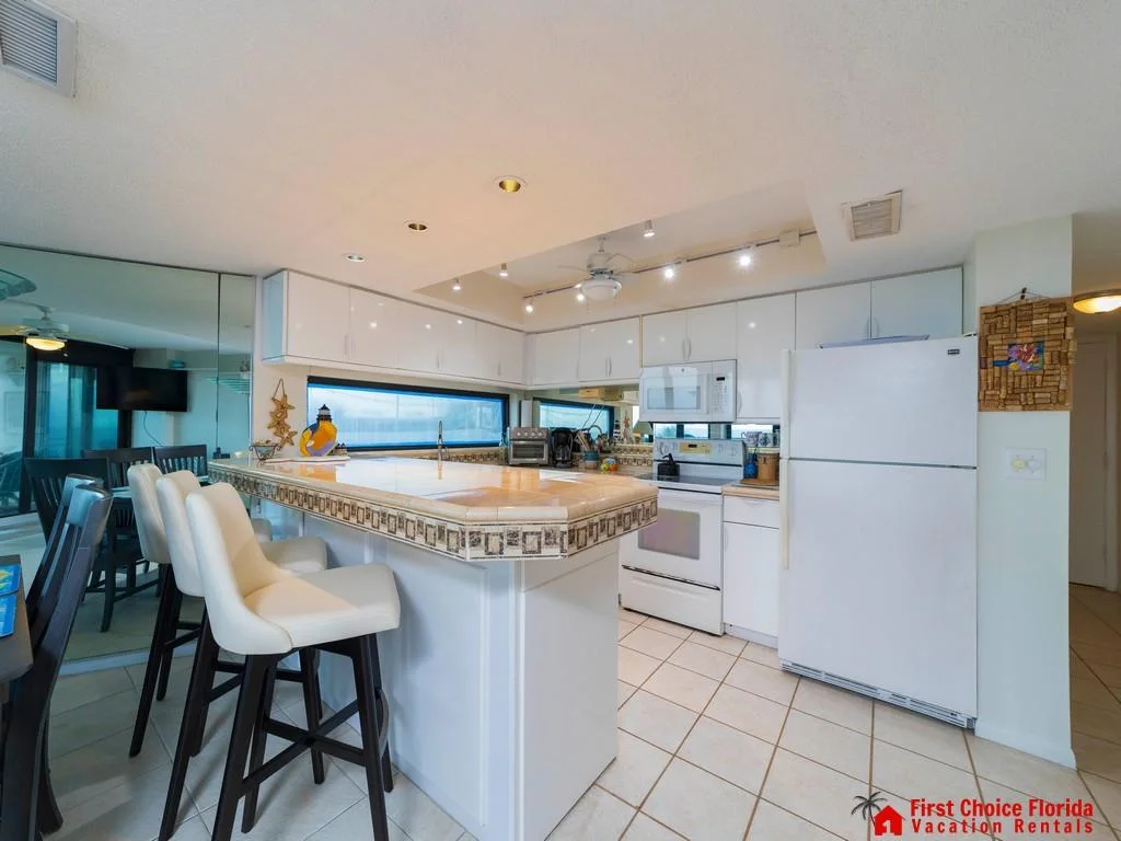 Breakfast bar with tile countertop and cushioned stools overlooks white cabinetry and appliances in the kitchen.