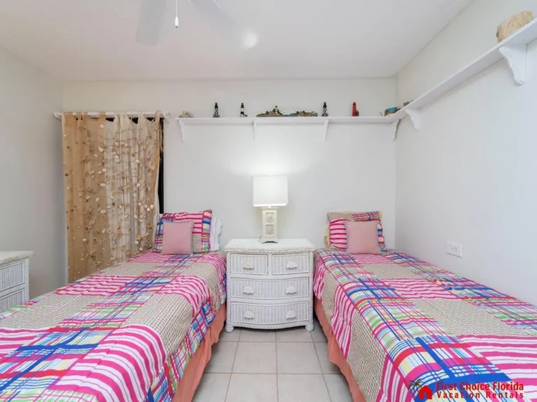 Twin beds with colorful plaid bedding flank a white wicker nightstand topped by a table lamp in a tiled bedroom.