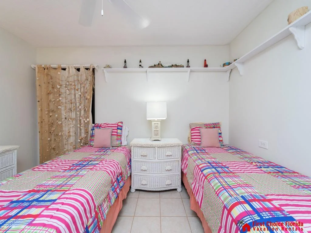 Twin beds with colorful plaid bedding flank a white wicker nightstand topped by a table lamp in a tiled bedroom.