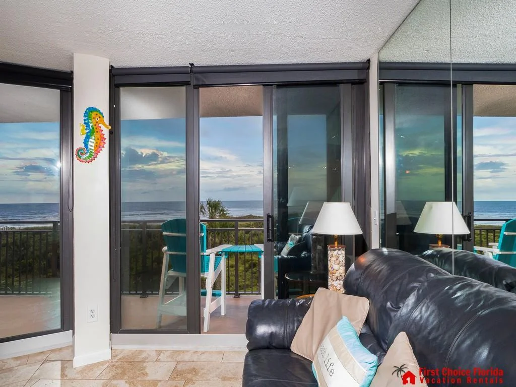 Black leather sofa with coastal pillows faces sliding glass doors opening to an oceanfront balcony with blue Adirondack chairs.