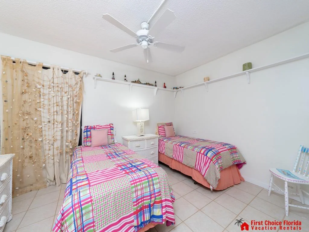 Twin beds with colorful patchwork quilts flank a white wicker nightstand beneath wall shelves and seashell curtains.