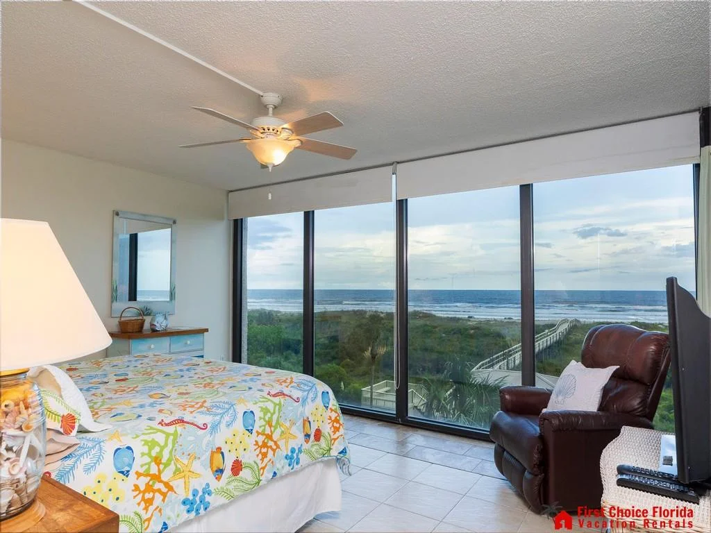 Ocean-view bedroom with tile flooring, ceiling fan, leather recliner, and colorful coral-patterned bedding.