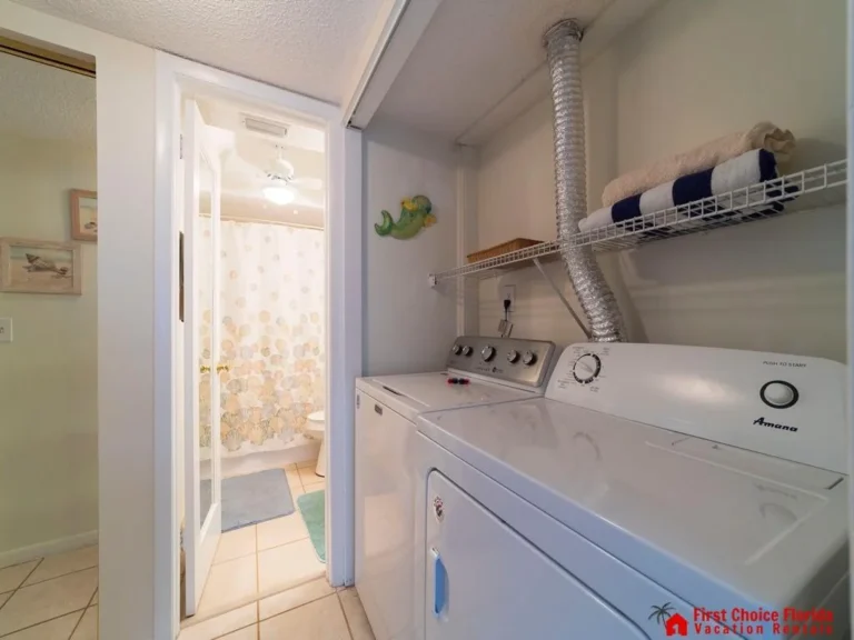 Laundry area with white washer and dryer beneath a wire shelf holding folded towels, next to a bathroom with seashell shower curtain.