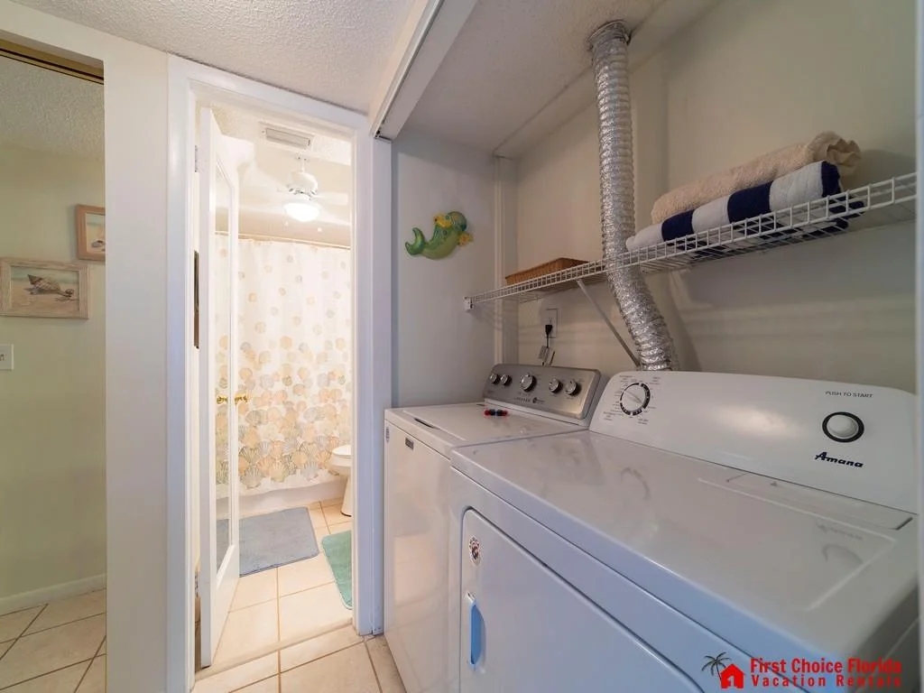 Laundry area with white washer and dryer beneath a wire shelf holding folded towels, next to a bathroom with seashell shower curtain.
