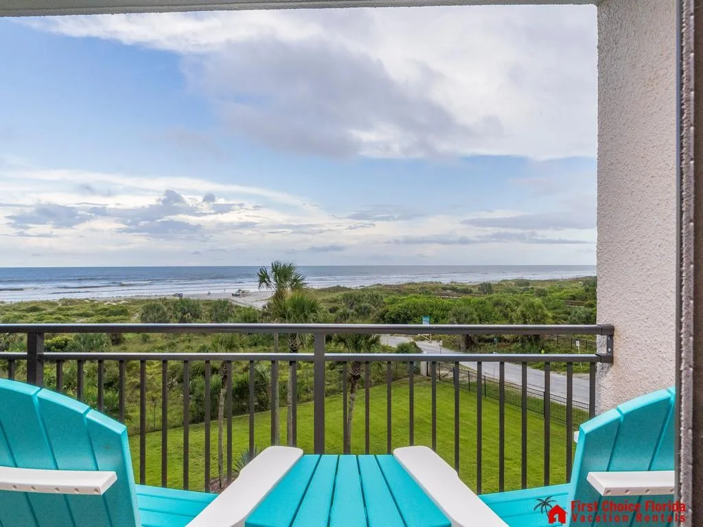 Clear blue Adirondack chairs face the ocean beyond a black metal balcony railing under a cloudy sky.