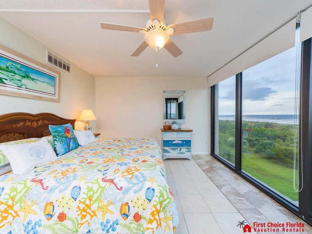 Tile flooring extends to floor-to-ceiling windows framing beach and ocean views beyond a painted dresser and ceiling fan.