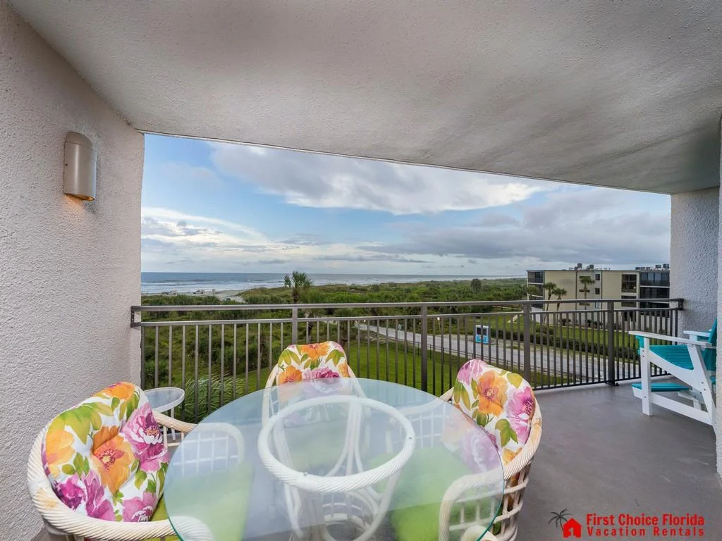 Covered balcony with glass-top table, floral cushioned chairs, and views of greenery and ocean beyond.