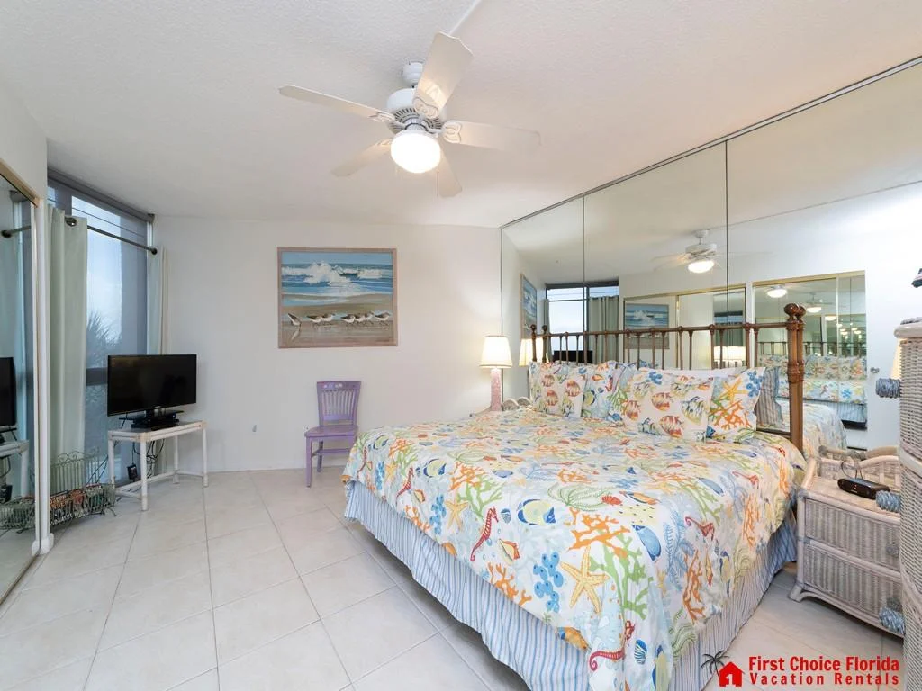 Mirrored wall expands the bedroom space with a ceiling fan above and a wicker nightstand beside the king bed.