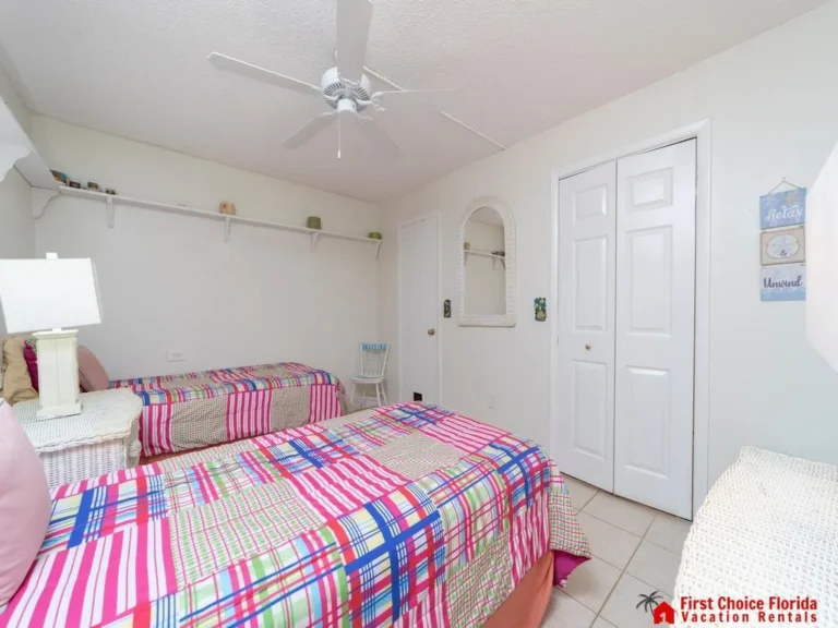 Twin beds with colorful plaid bedding face a white double-door closet in a tiled bedroom with ceiling fan.