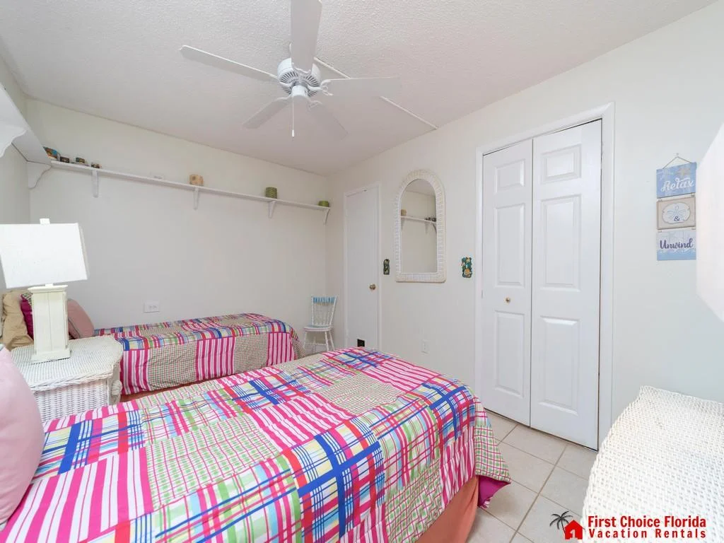 Twin beds with colorful plaid bedding face a white double-door closet in a tiled bedroom with ceiling fan.