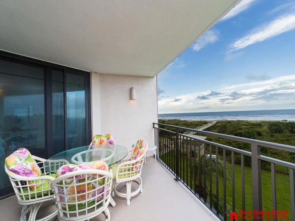 Oceanfront balcony features a glass-top table with floral cushioned chairs and views of the beach walkway.
