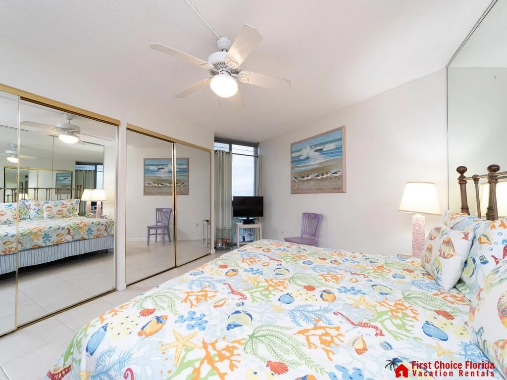 Bedroom with mirrored sliding closets, ceiling fan, colorful ocean-themed bedding, and a TV on a white stand.