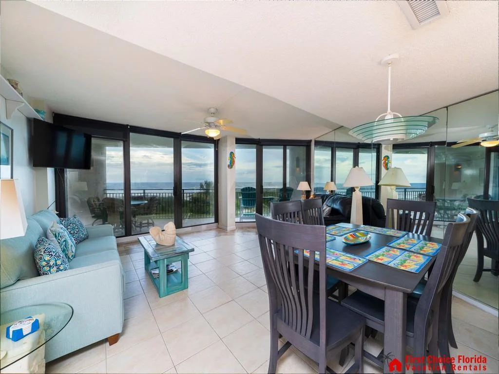 Dining table with colorful placemats sits beside a mirrored wall reflecting black leather sofa and ocean views.