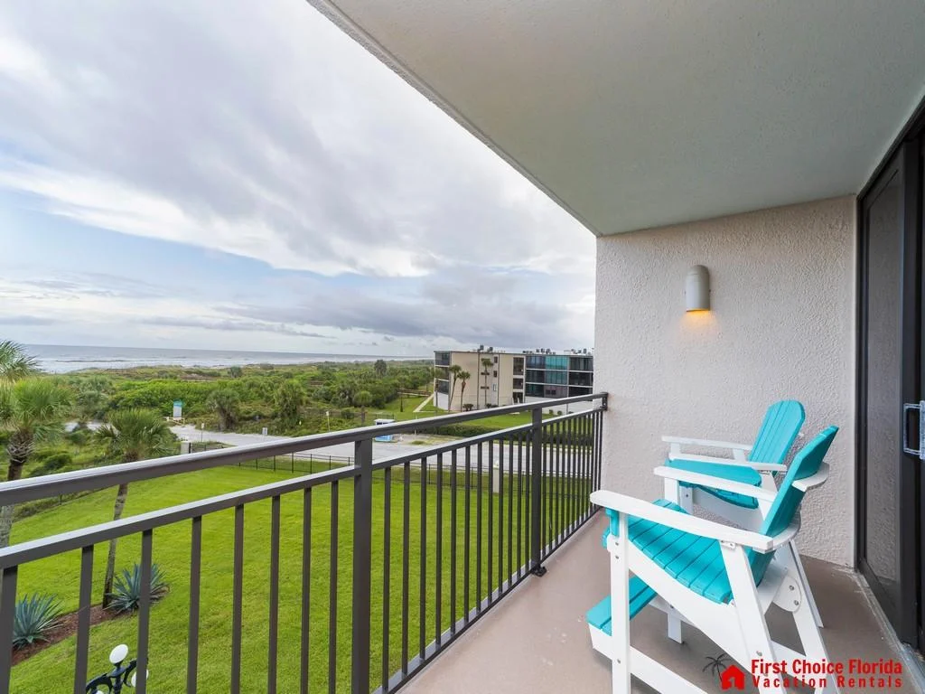Oceanfront balcony with turquoise Adirondack chairs, black metal railing, and views of greenery and ocean horizon.