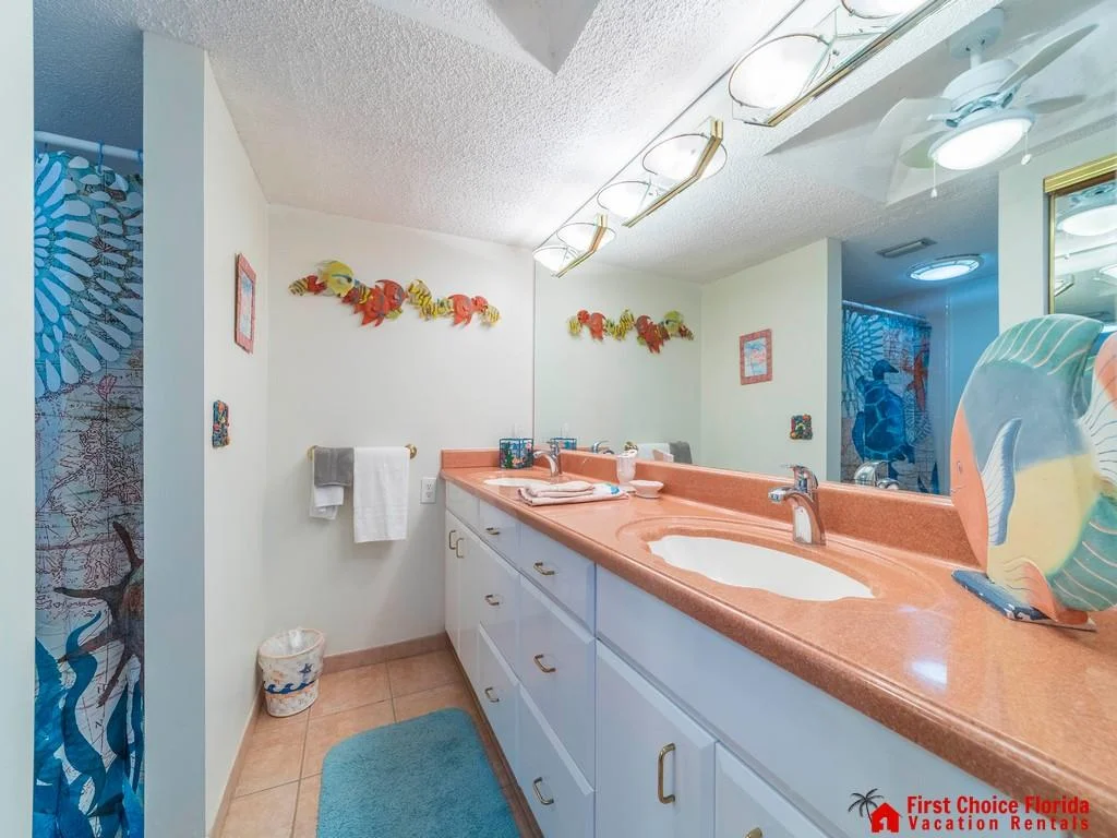 Long coral-colored countertop with dual sinks stretches beneath a wide mirror and brass light fixtures.
