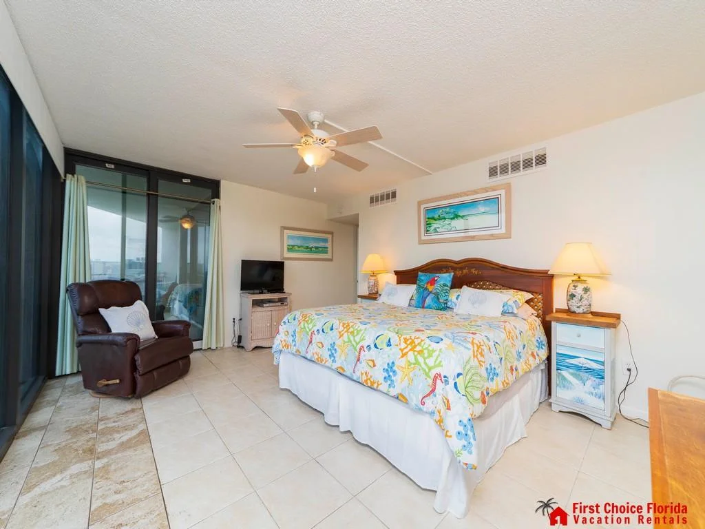 King bed with colorful coral and sea life bedding faces a TV on a wicker stand, next to a leather recliner.