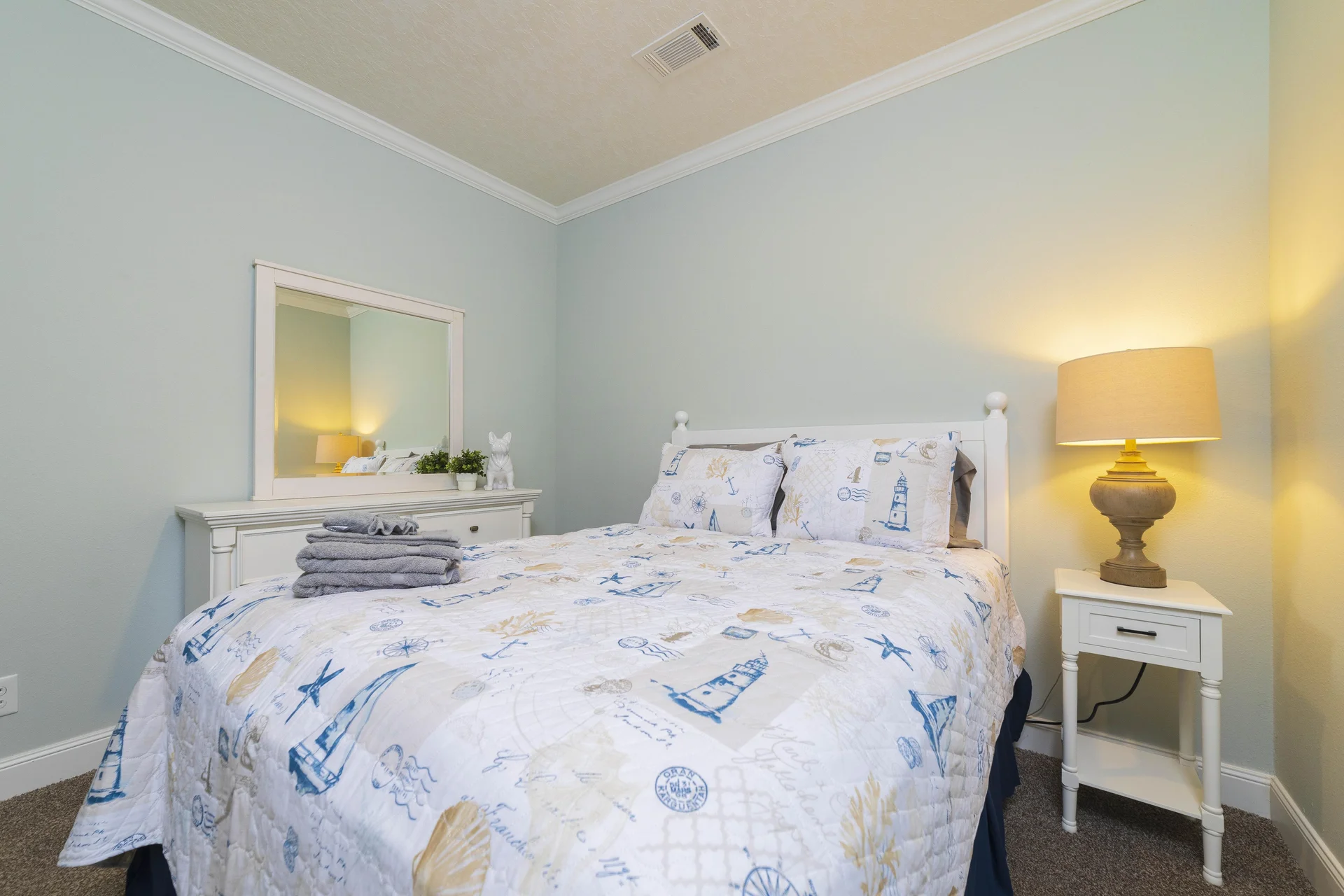 Stack of folded gray towels rests on a nautical-themed bedspread with a beige table lamp glowing on a white nightstand.