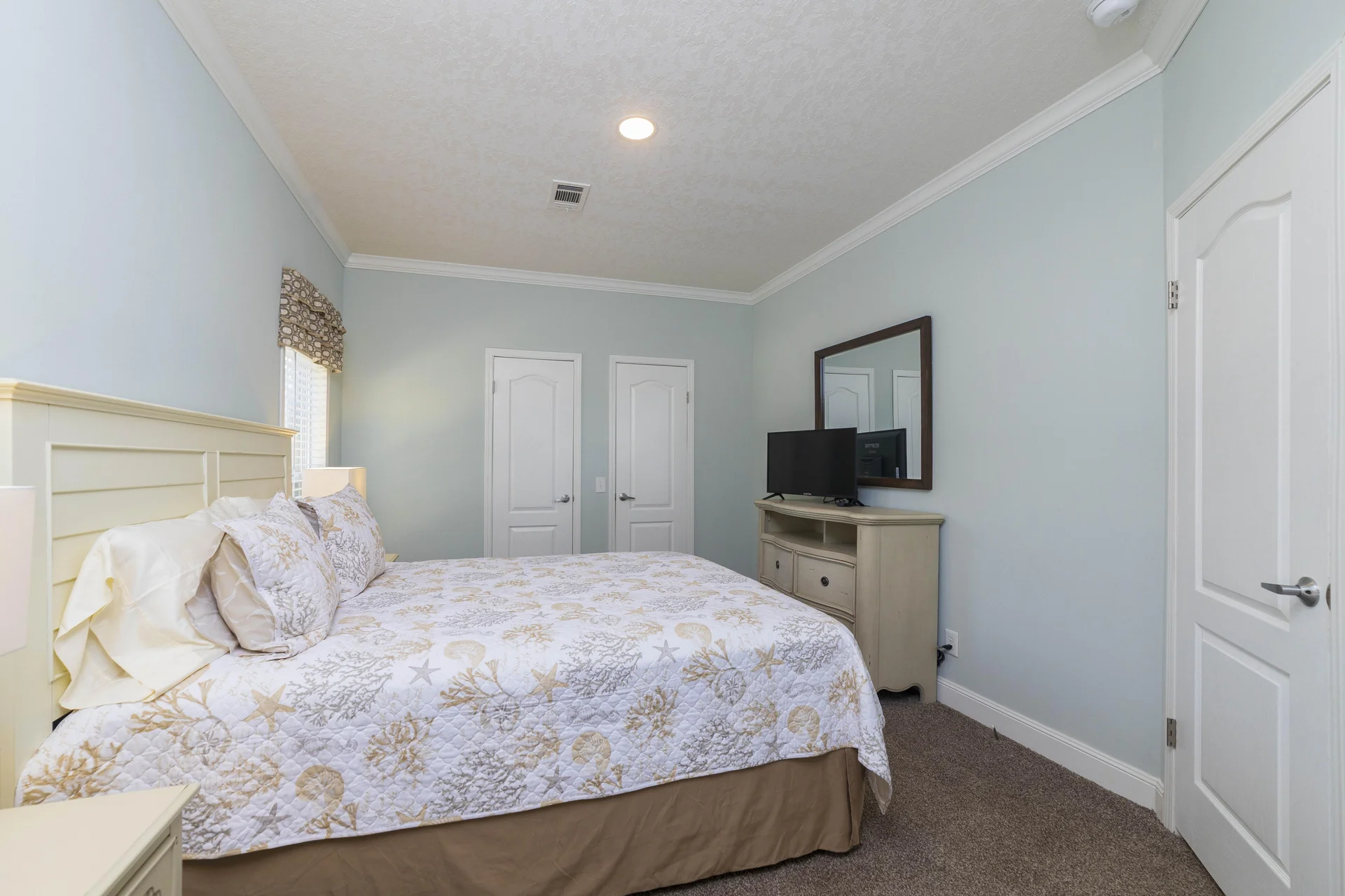 Bedroom with light blue walls, patterned bedspread, beige carpet, and TV on a cream dresser beneath a large mirror.