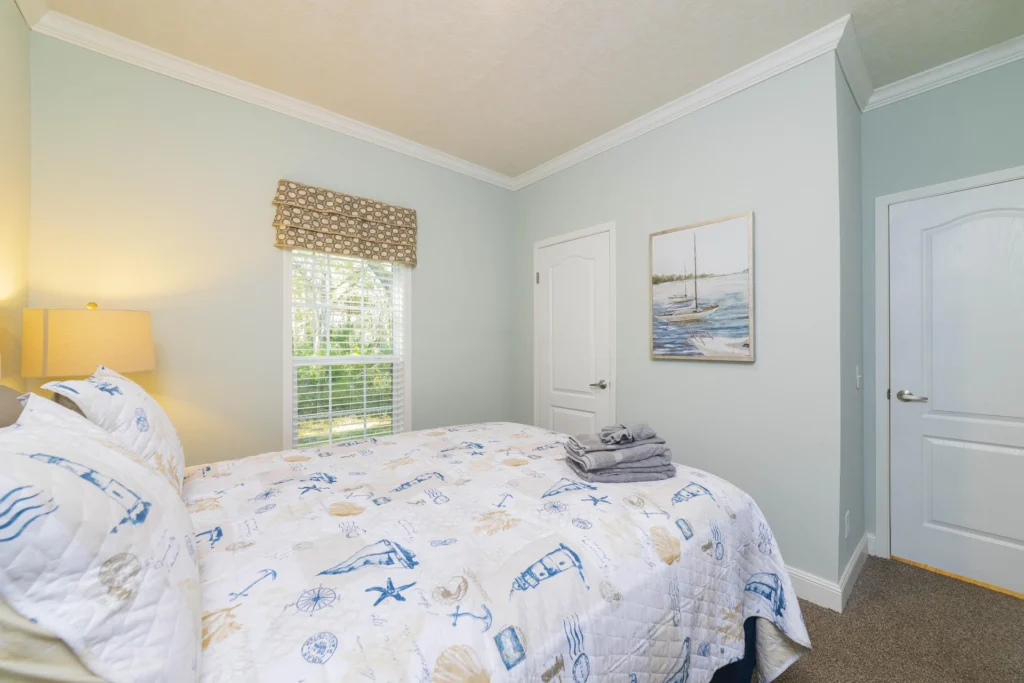 Queen bed with nautical-themed quilt and pillows, beige carpet, light blue walls, window with patterned roman shade.