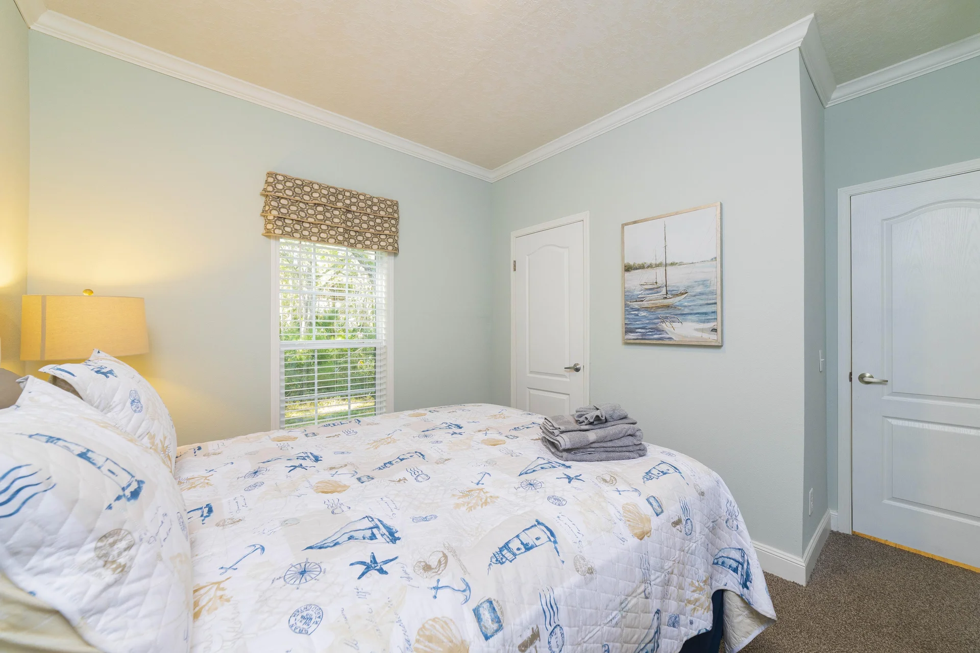 Queen bed with nautical-themed quilt and pillows, beige carpet, light blue walls, window with patterned roman shade.