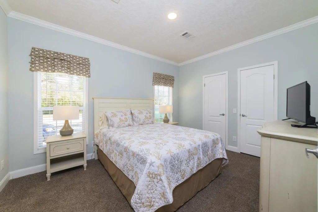 Two windows with patterned roman shades flank a queen bed featuring a coastal-themed quilt and matching pillows.