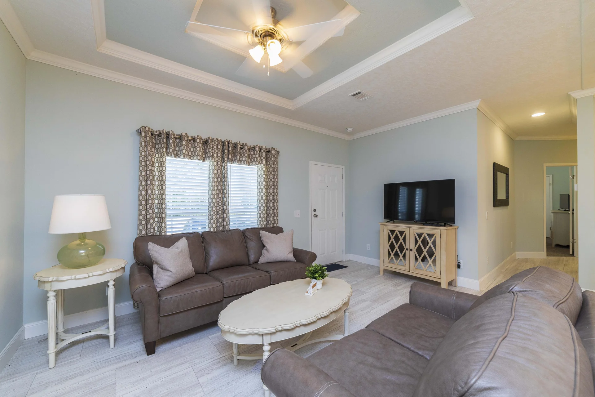 Living room with gray leather sofas, oval cream coffee table, patterned curtains, and ceiling fan with lights.