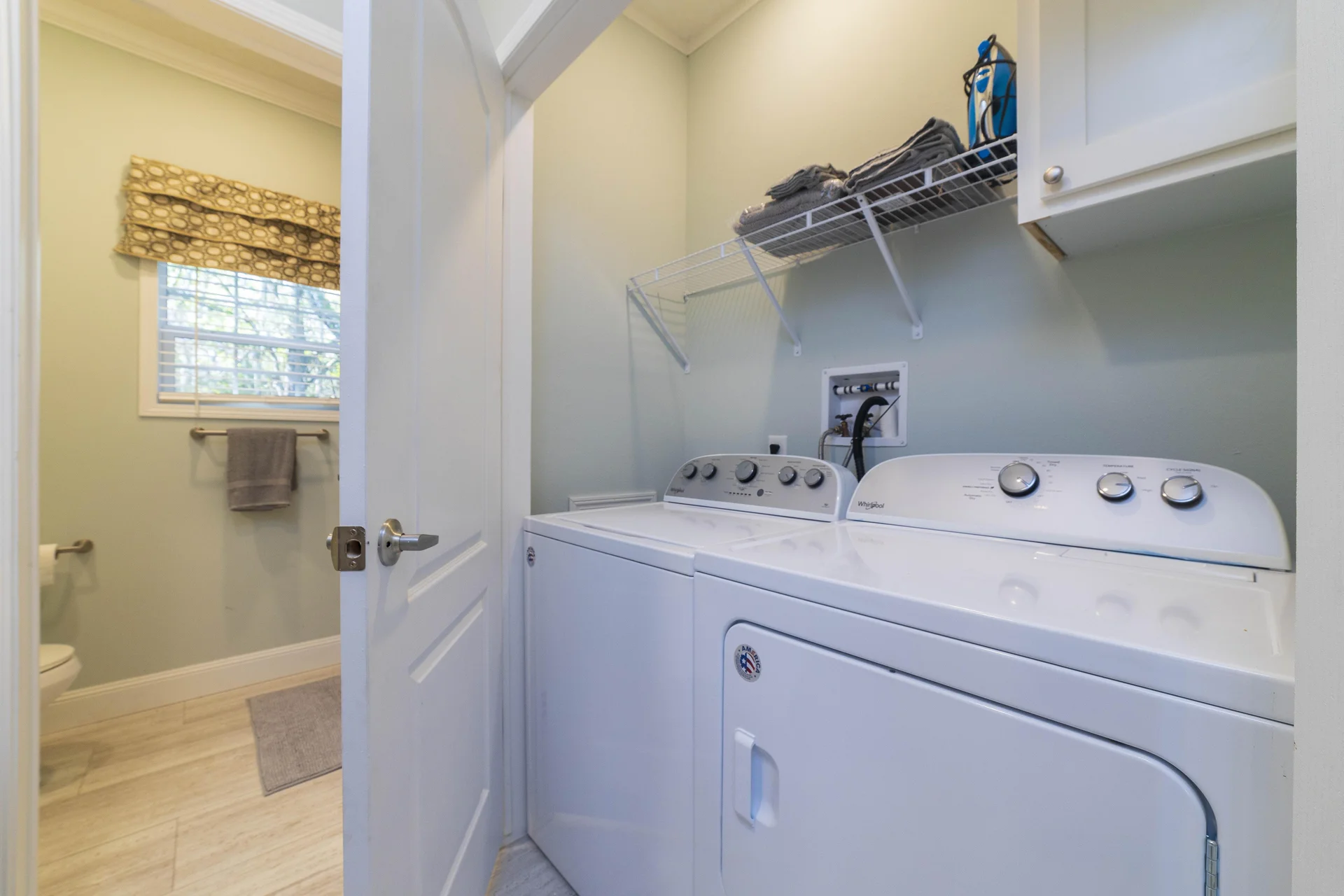 Laundry room with white washer and dryer beneath a wire shelf holding folded towels and an iron.