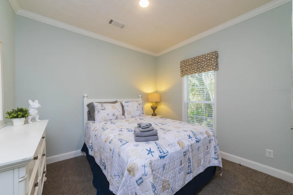 Window with white blinds and patterned roman shade beside a bed covered in a nautical quilt and stacked towels.
