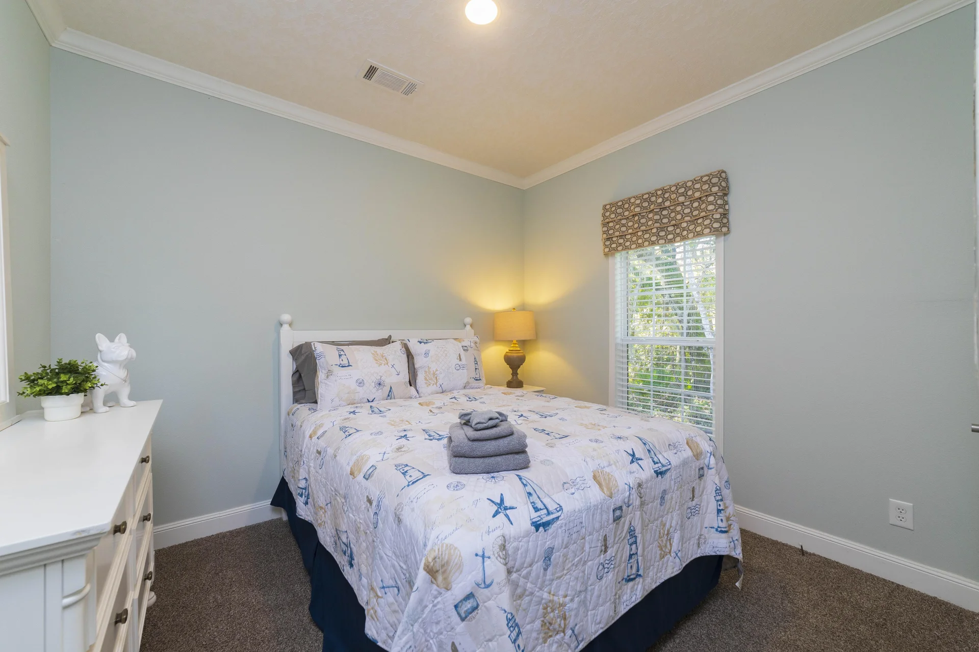 Window with white blinds and patterned roman shade beside a bed covered in a nautical quilt and stacked towels.