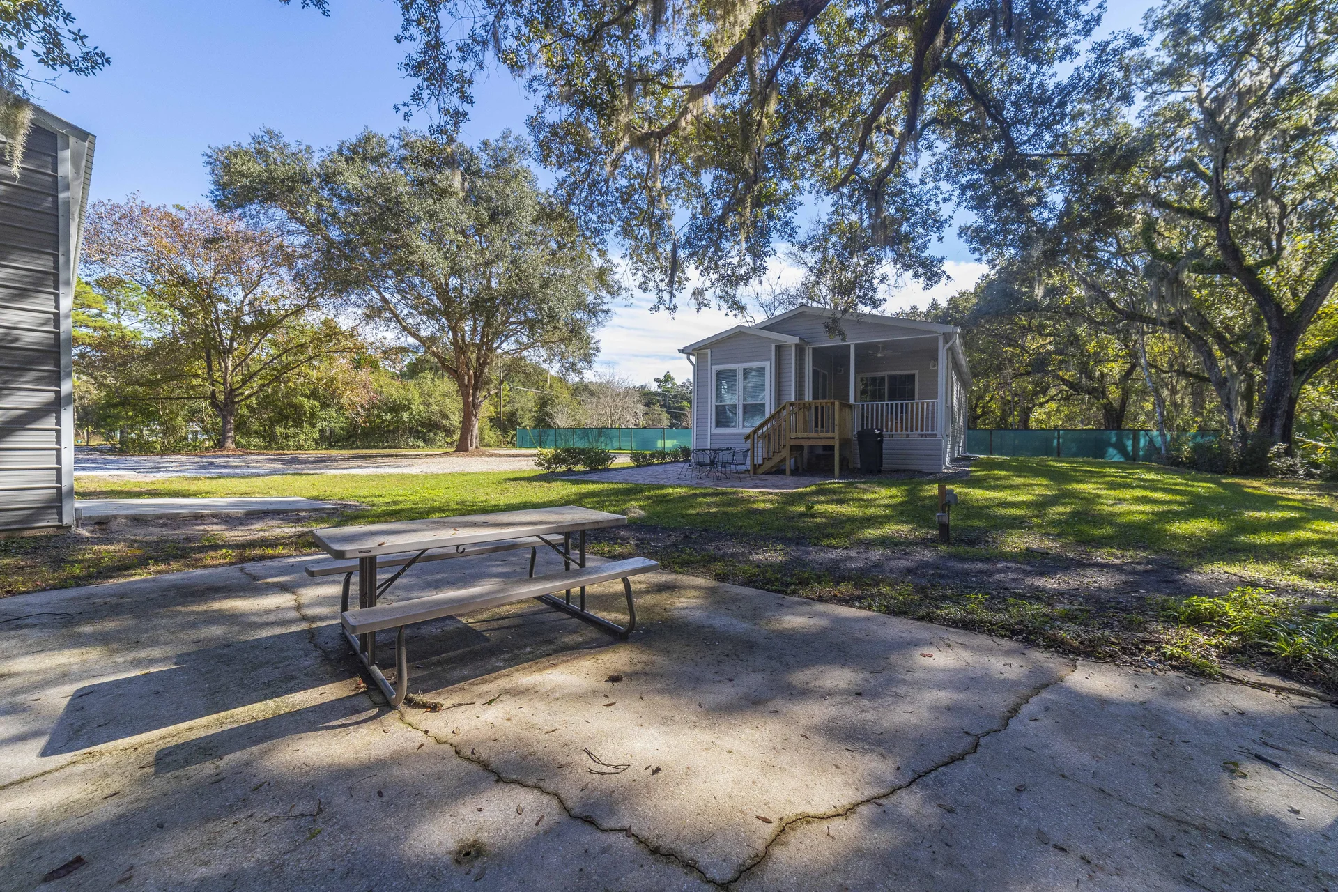 Concrete patio with a wooden picnic table set beneath sprawling oak trees and a vinyl-sided house in the background.