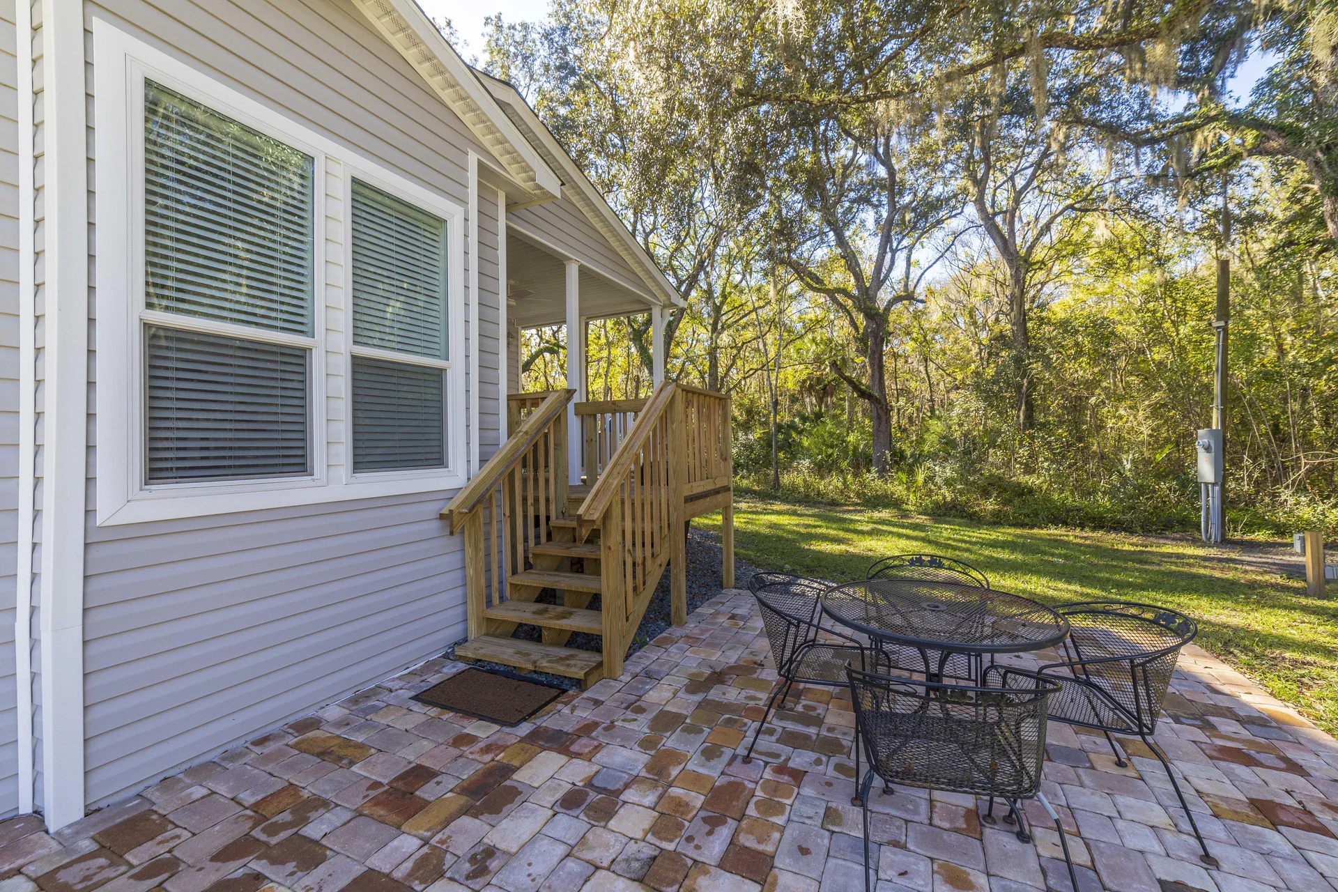 Brick patio with black metal dining set beside a beige vinyl house exterior and wooden steps leading to a covered porch.