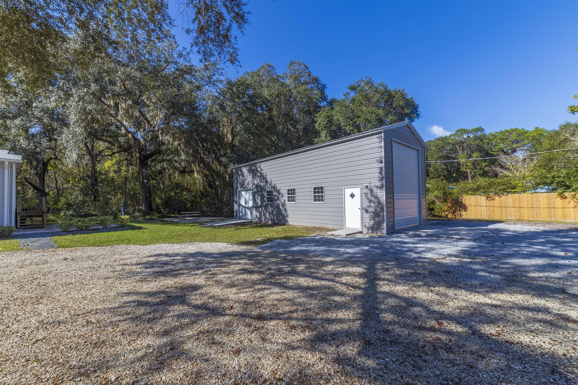 Gravel driveway and open yard shaded by oak trees, with a large gray metal garage and wooden fence.