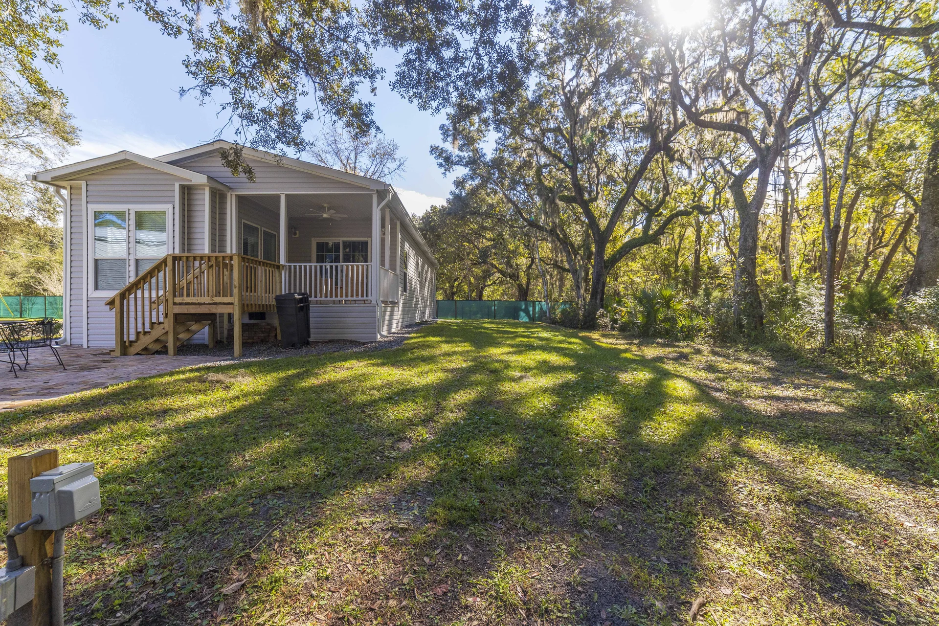 Side yard featuring sun-dappled grass, mature oak trees, and a fenced backyard adjacent to the house’s covered porch.