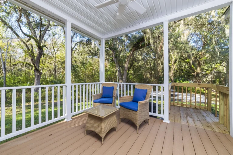 Screened porch with wicker chairs featuring blue cushions and a matching glass-top table, overlooking oak trees.